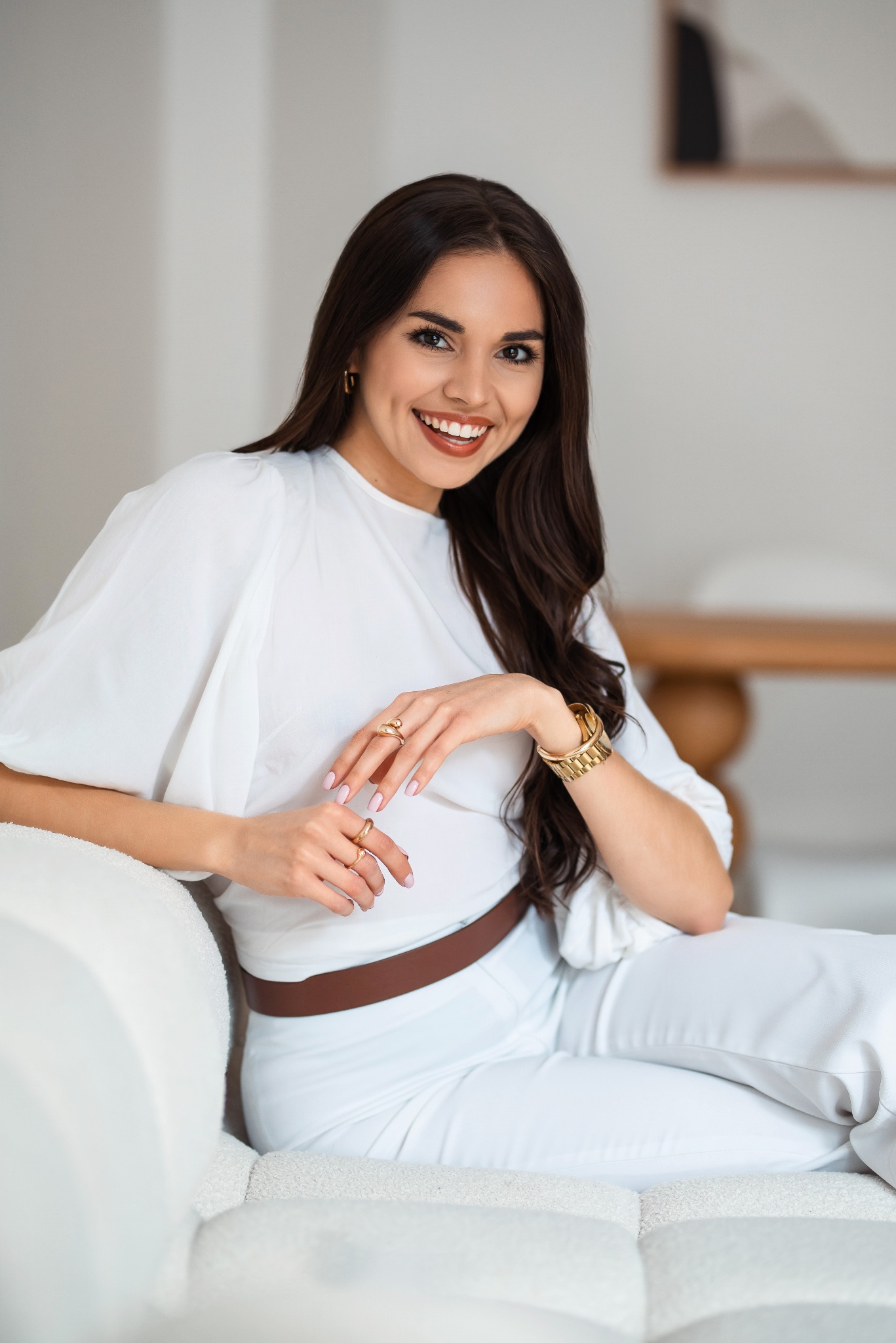 Uśmiechnięta brunetka w bieli siedzi na kanapie, prezentując złotą biżuterię: pierścionki i zegarek. Stonowane tło, naturalne światło. Elegancki, swobodny styl.
