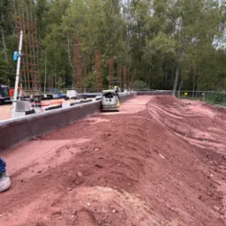 Praca ziemne: zagęszczarka wibruje czerwoną ziemię obok fundamentów z izolacją, w tle las i zbrojenia pod konstrukcje. Widoczne ślady opon i butów.