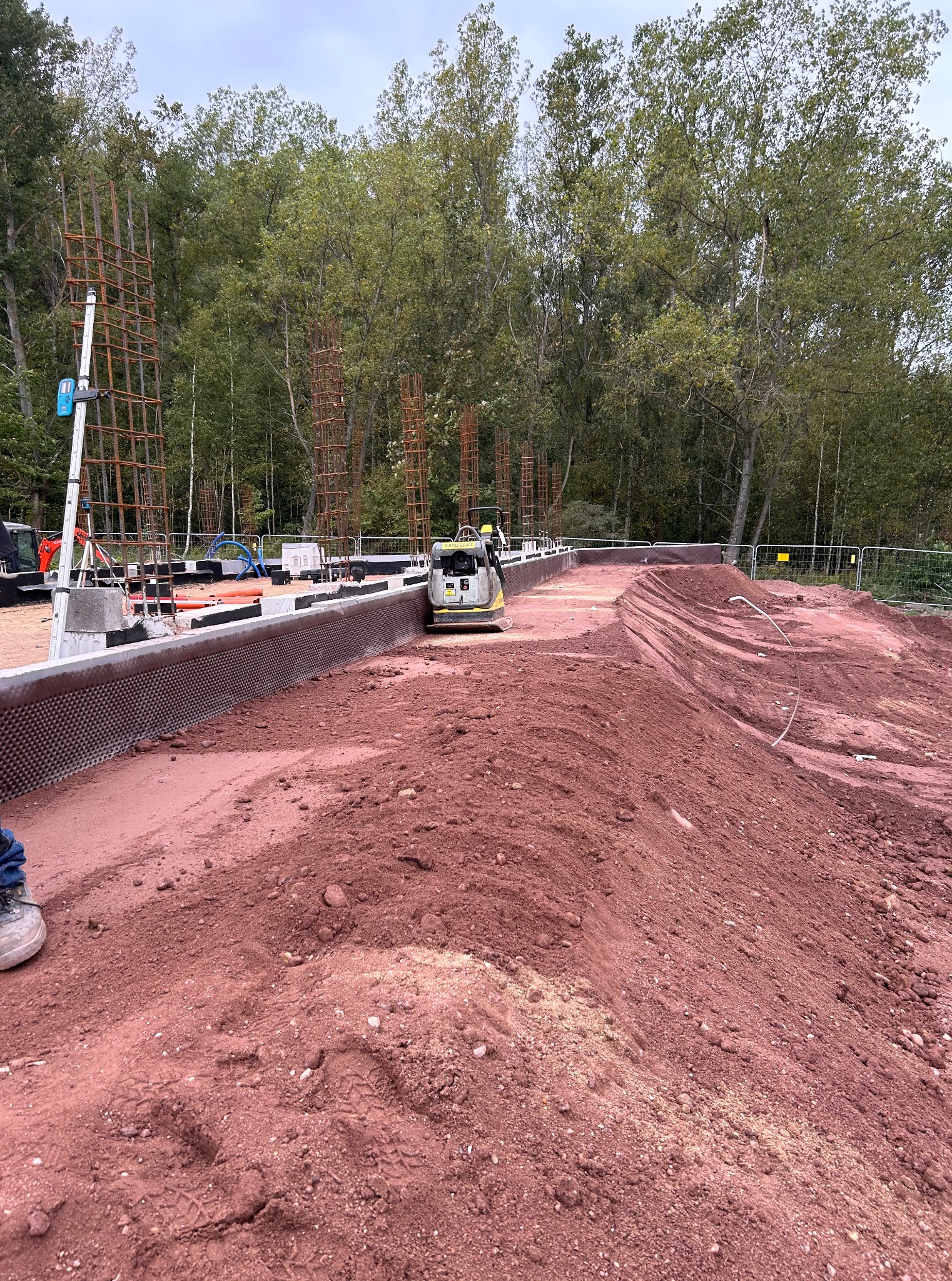 Praca ziemne: zagęszczarka wibruje czerwoną ziemię obok fundamentów z izolacją, w tle las i zbrojenia pod konstrukcje. Widoczne ślady opon i butów.
