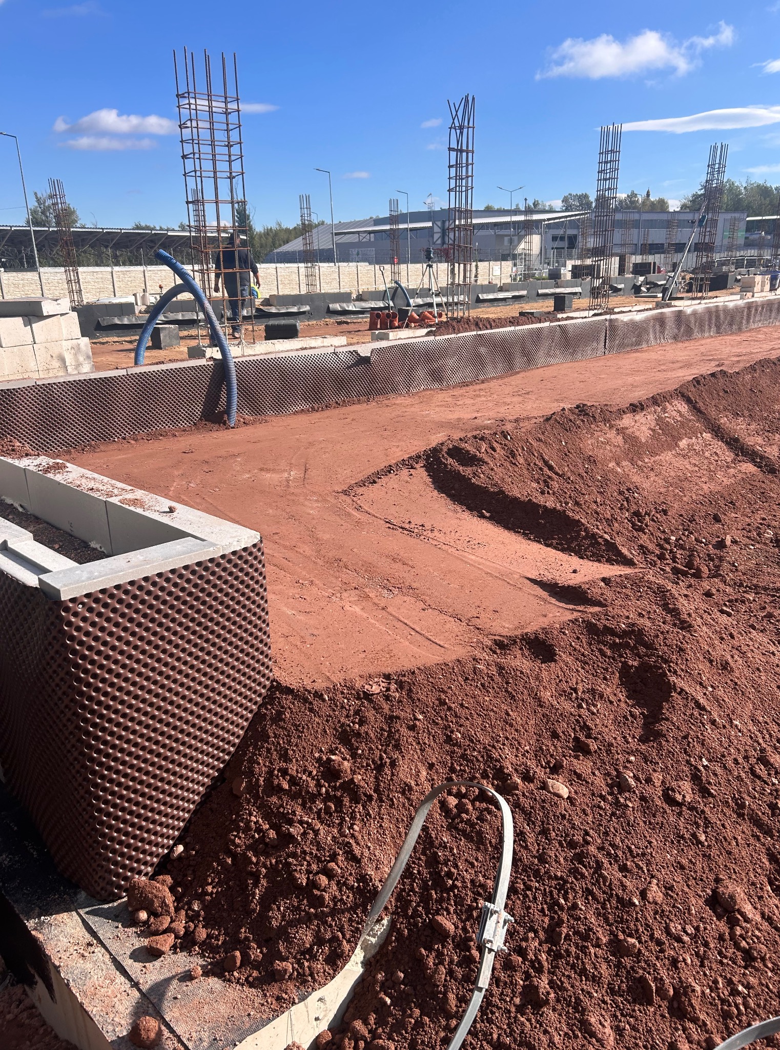 Fundamenty budynku w trakcie budowy, widoczne zbrojenia, bloczki betonowe i czerwona ziemia. W tle budynek i błękitne niebo.