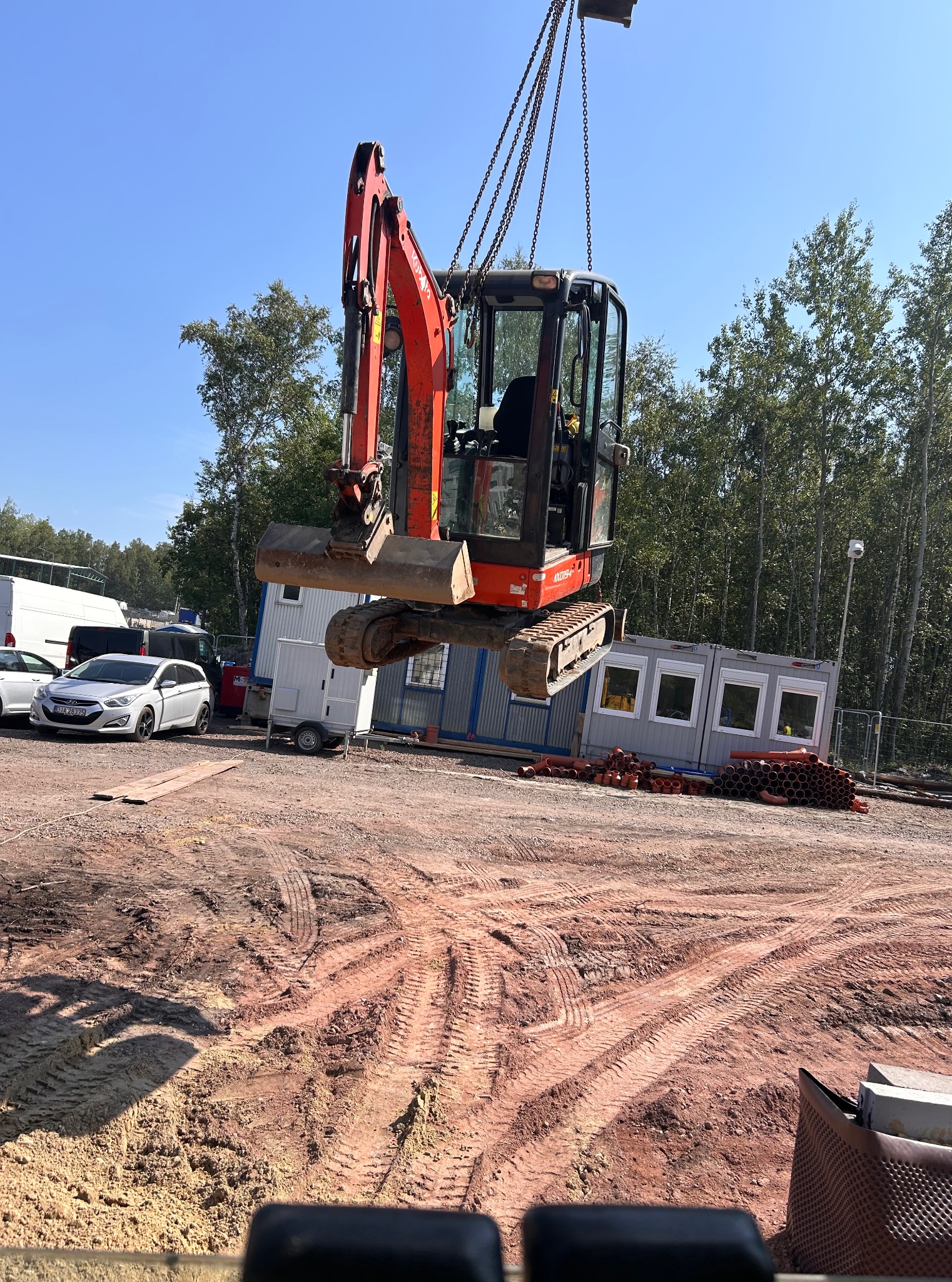 Podwieszana, czerwona minikoparka Kubota na łańcuchach, unoszona nad placem budowy z widocznymi śladami opon i rurami PCV w tle. Transport koparki na budowie.