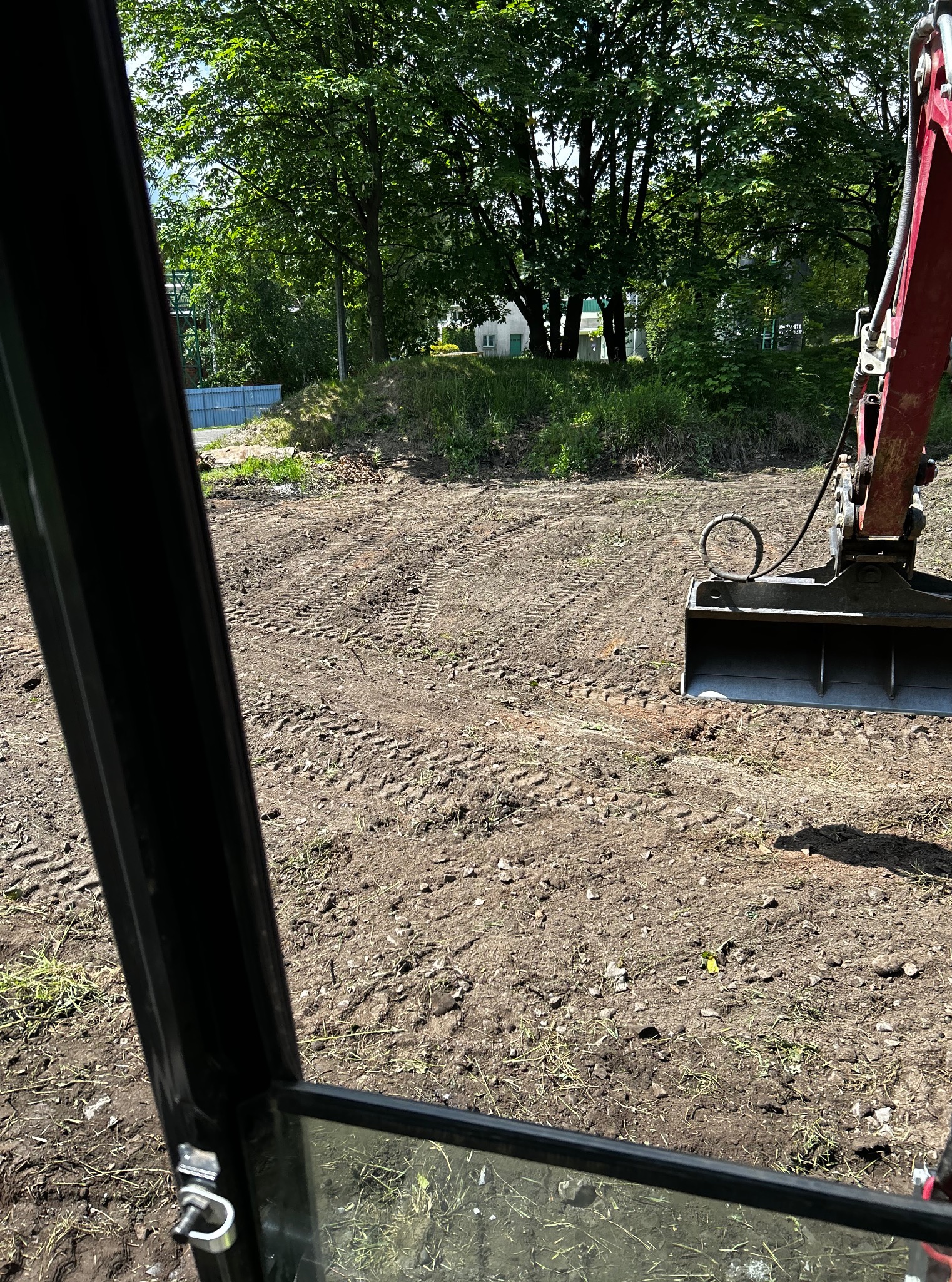 Widok z kabiny koparki na wyrównany teren z widocznymi śladami opon, w tle zieleń drzew i budynek.