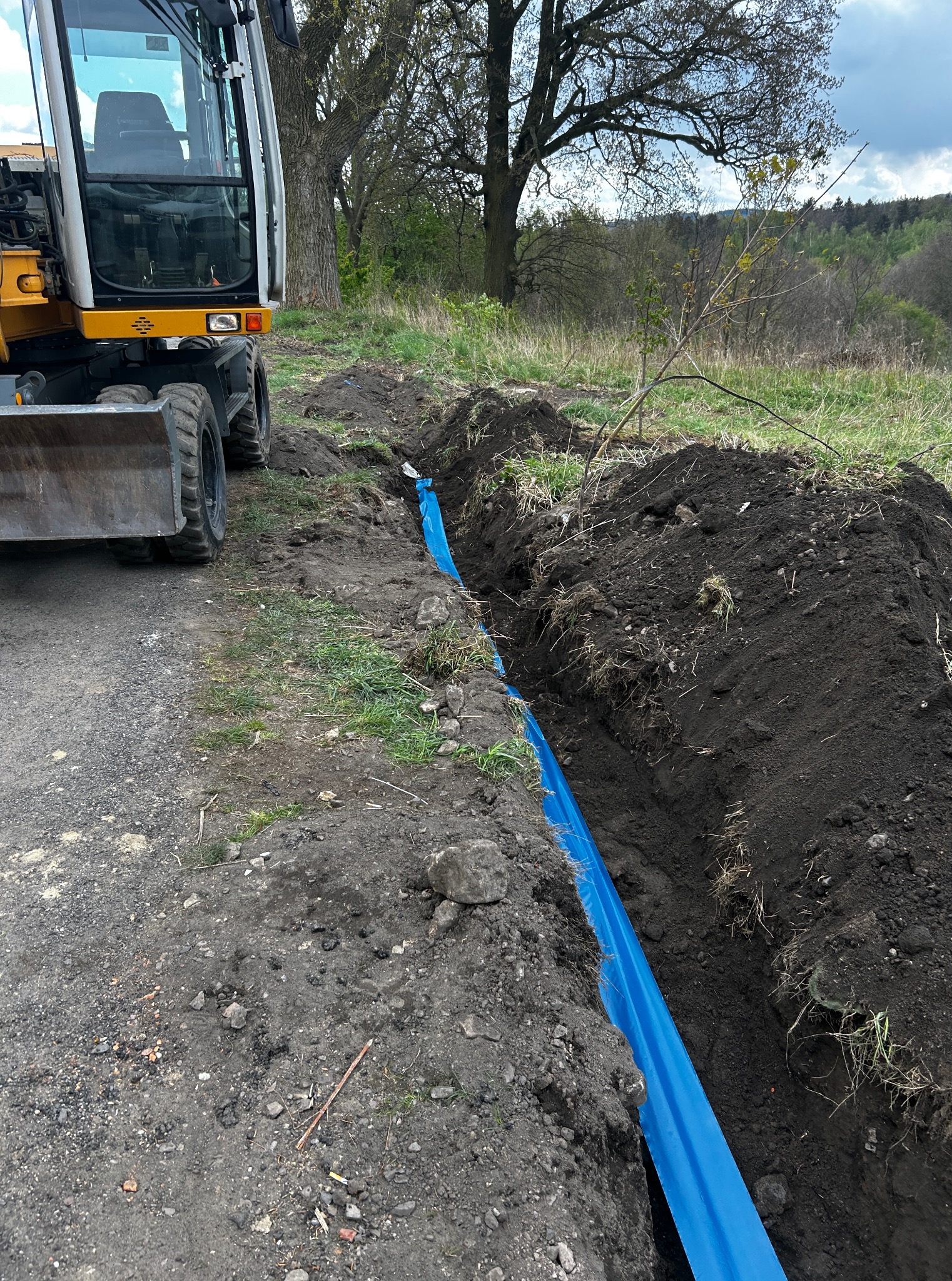 Żółta minikoparka Liebherr obok wykopu zabezpieczonego niebieską folią, widok na teren zielony w tle. Etap prac ziemnych w terenie niezabudowanym.