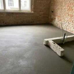 Świeżo wylana, gładka posadzka jastrychowa w surowym wnętrzu z ceglanymi ścianami i oknem. Widoczne narzędzia i rury wystające z podłoża.