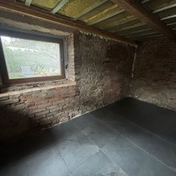 Demolka - Rozbiórki, Wyburzenia, Demontaże, Remonty Jelenia Góra - Wnętrze w trakcie remontu: surowe ceglane ściany, nowe ciemne płytki podłogowe, okno z widokiem na zieleń i izolacja stropu z metalowymi profilami.