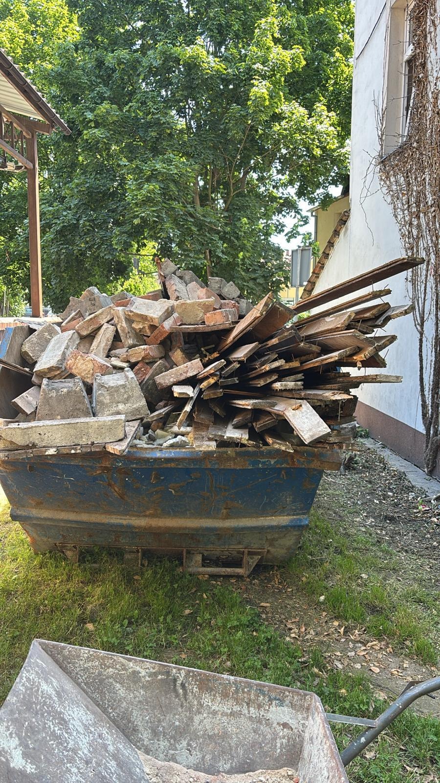 Kontener na gruz wypełniony cegłami, betonem i drewnem na trawniku, w tle budynek z bluszczem i drzewa. W rogu widoczna taczka.