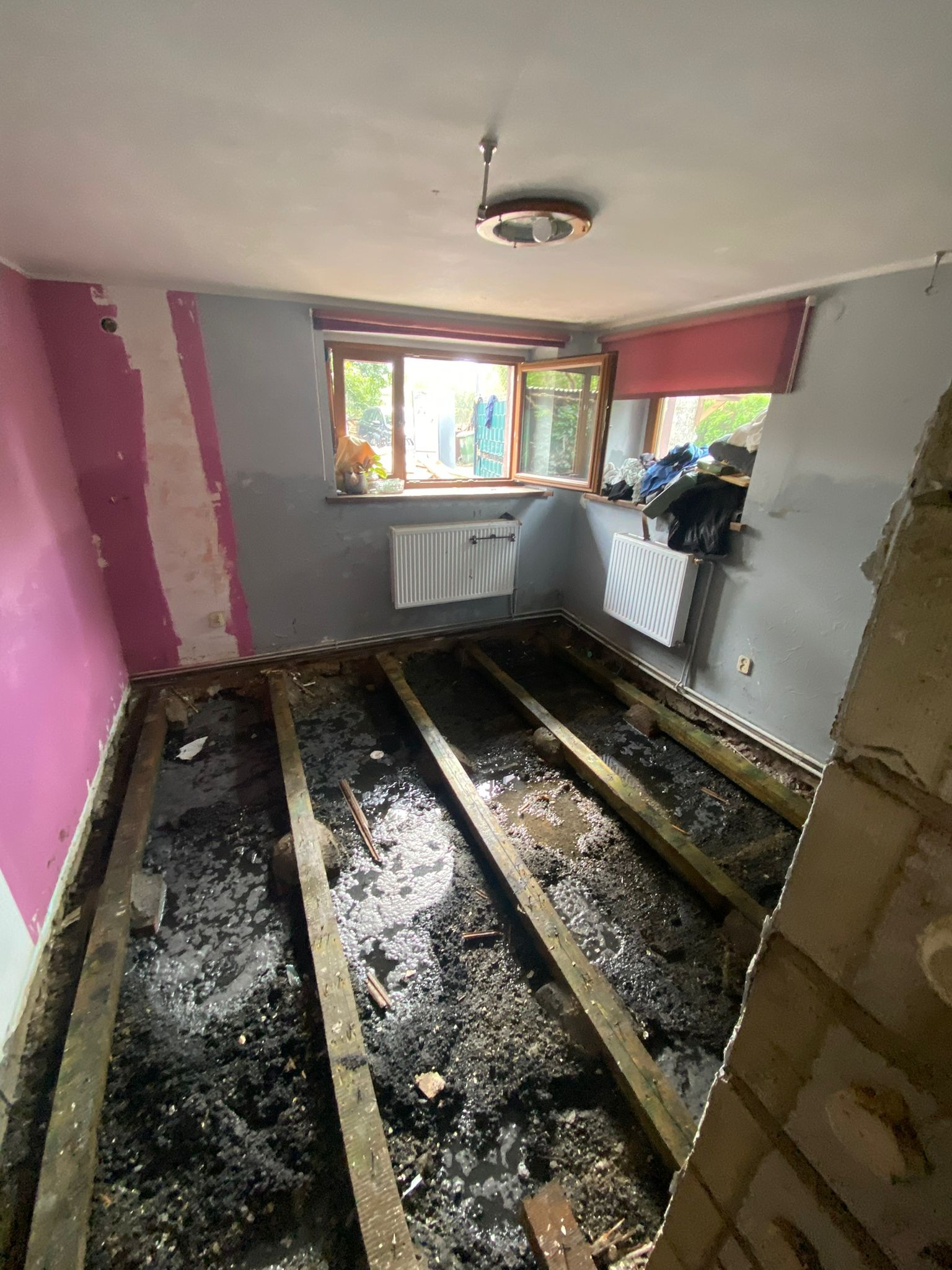 Pokój w trakcie remontu: usunięta podłoga, odsłonięte legary, ściany częściowo pomalowane, widoczne grzejniki i okno. Ukazany stan przygotowania do dalszych prac.