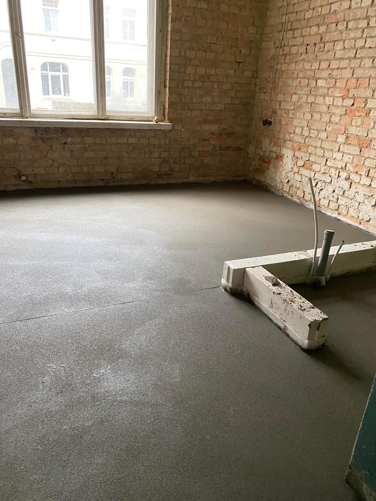 Świeżo wylana, gładka posadzka jastrychowa w surowym wnętrzu z ceglanymi ścianami i oknem. Widoczne narzędzia i rury wystające z podłoża.