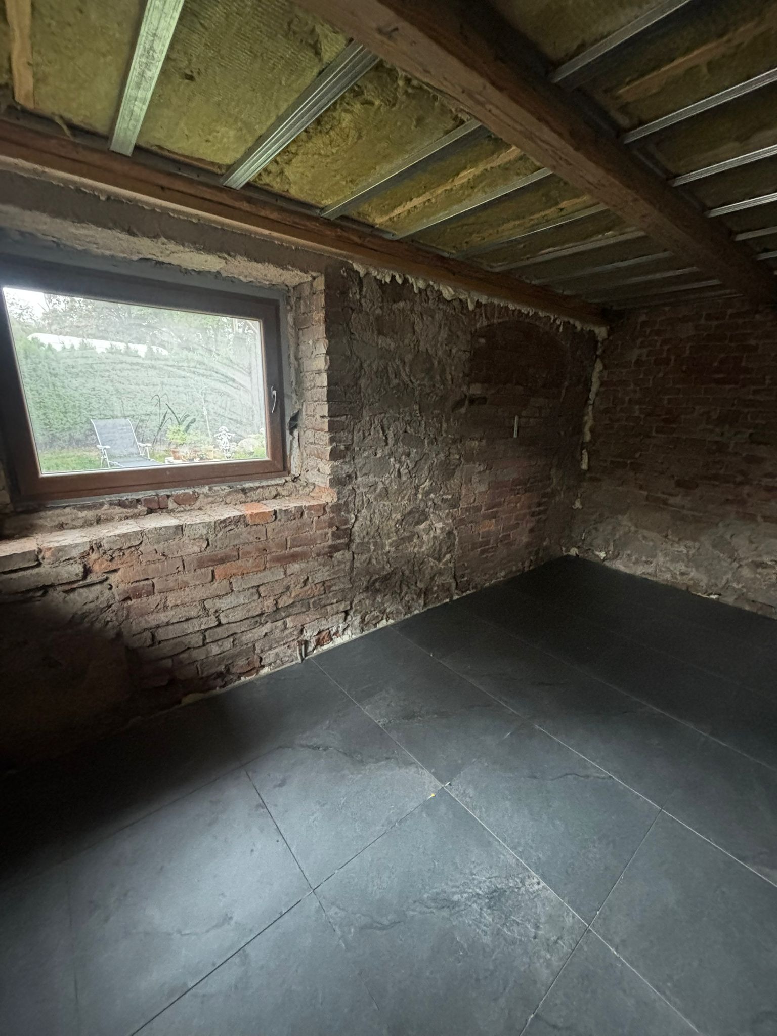 Wnętrze w trakcie remontu: surowe ceglane ściany, nowe ciemne płytki podłogowe, okno z widokiem na zieleń i izolacja stropu z metalowymi profilami.