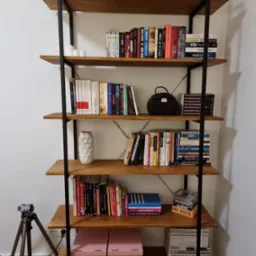 Ręcznie robiony regał z drewna i metalu, z książkami, dekoracjami i akcesoriami, ustawiony na tle białej ściany. Widoczny statyw kamery w lewym dolnym rogu.