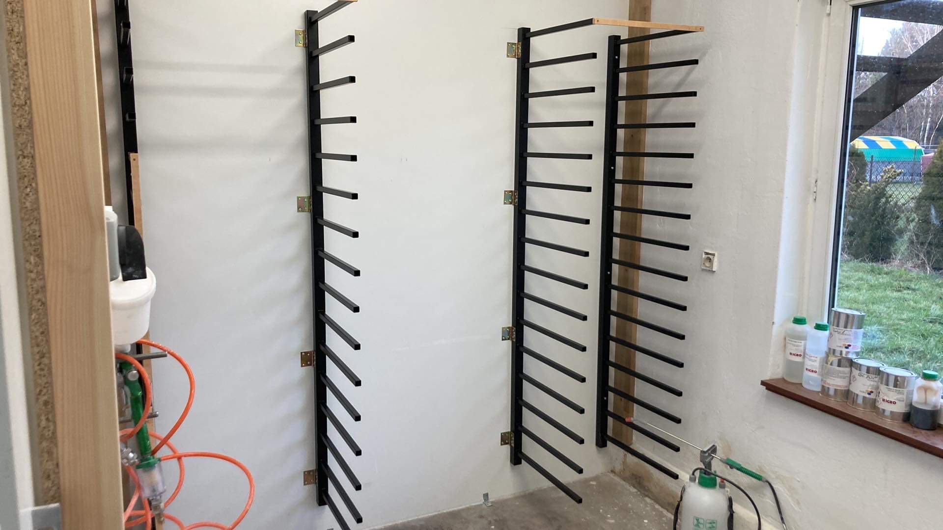 Metalowe, czarne konstrukcje regałowe zamontowane na białej ścianie w pomieszczeniu warsztatowym, widok z przodu, z oknem i narzędziami w tle.