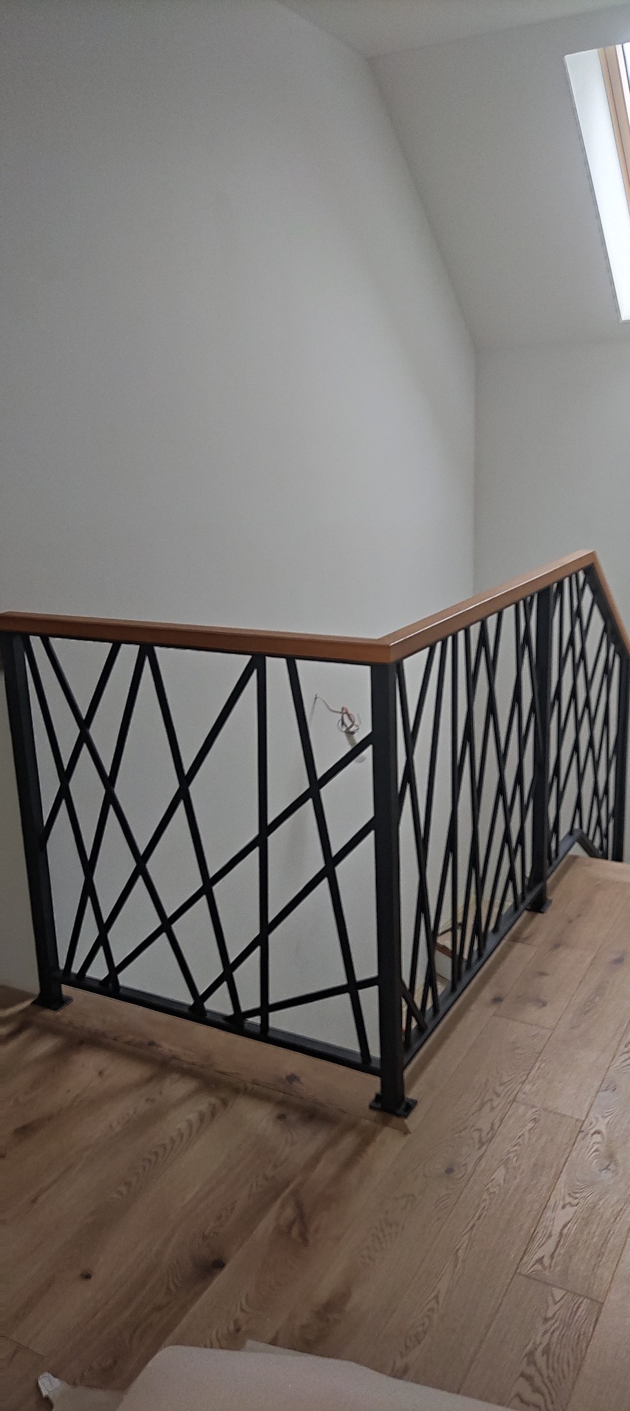 Czarna balustrada schodowa z drewnianym poręczem, o geometrycznym wzorze, na tle białej ściany i drewnianej podłogi. Widok z dołu ukazuje detale wykonania.