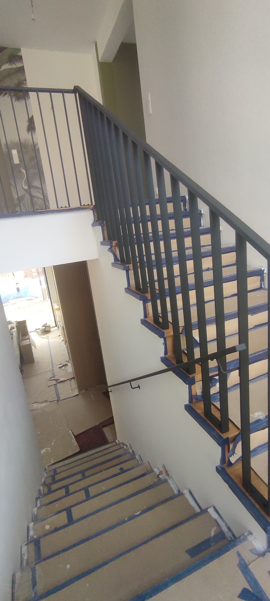 Ujęcie z góry na schody z drewnianymi stopniami zabezpieczonymi taśmą malarską i ciemnymi, pionowymi balustradami. Widoczne zabezpieczenia podłogi kartonem.