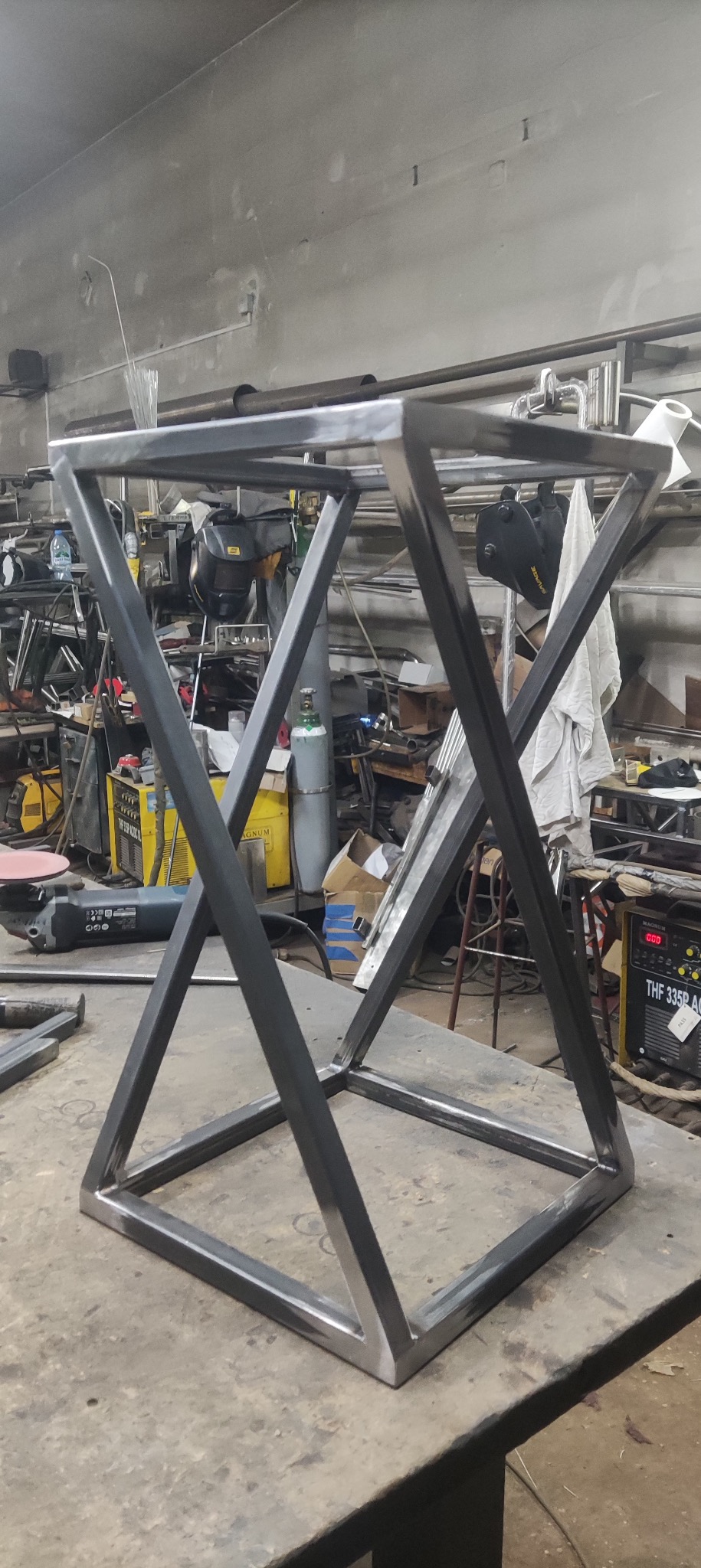 Metalowa konstrukcja geometryczna w warsztacie, spawana z profili kwadratowych, surowy wygląd, widoczne ślady obróbki, maszyna spawalnicza w tle.