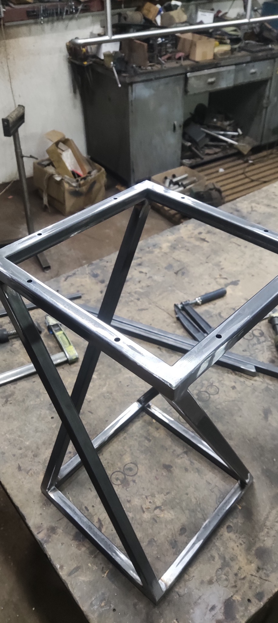 Metalowa konstrukcja w kształcie sześcianu z otworami, spawana, w warsztacie. Widoczne narzędzia i elementy obróbki metalu. Surowy, industrialny charakter.