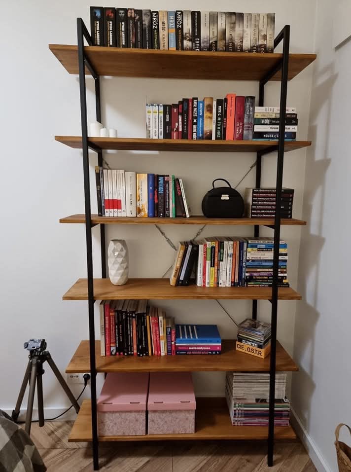 Ręcznie robiony regał z drewna i metalu, z książkami, dekoracjami i akcesoriami, ustawiony na tle białej ściany. Widoczny statyw kamery w lewym dolnym rogu.