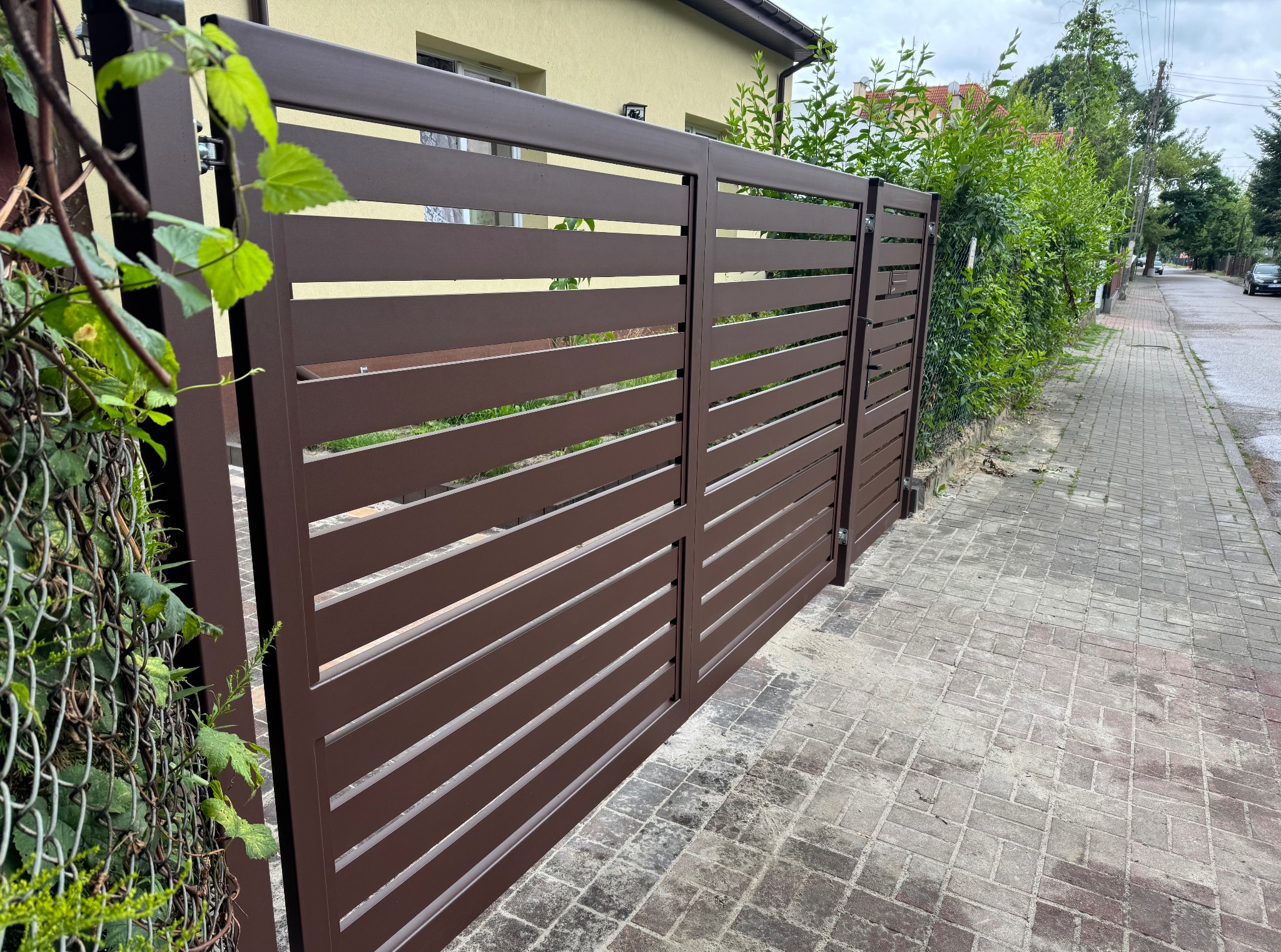 Nowoczesna brama wjazdowa z poziomymi lamelami w kolorze brązowym, widoczna od strony ulicy. Brama zamontowana przy posesji z kostką brukową.