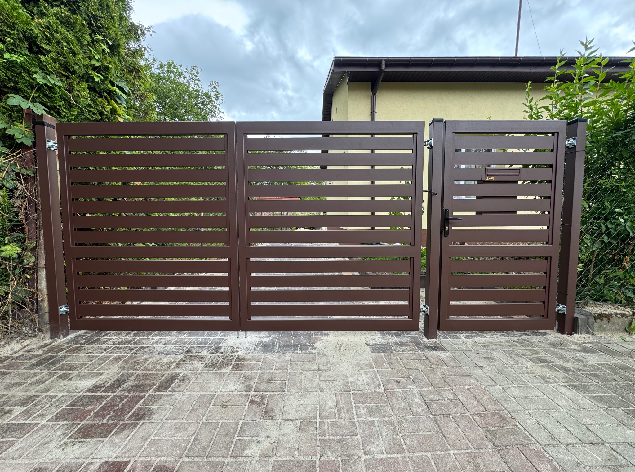 Nowoczesna, brązowa brama wjazdowa z poziomymi szczeblami, zintegrowana furtka z zamkiem i skrzynką pocztową. Kostka brukowa na podjeździe, zieleń w tle.
