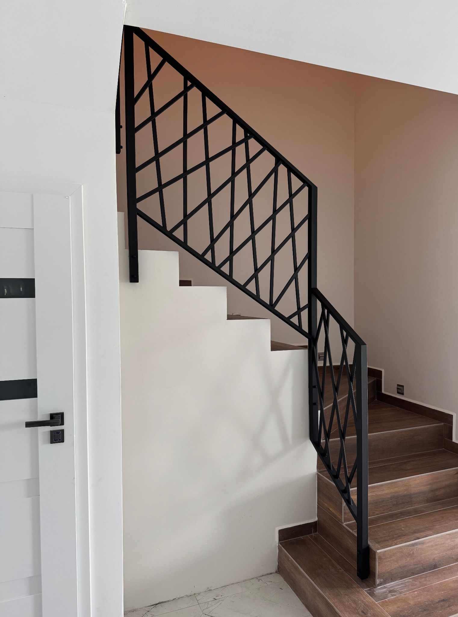 Minimalistyczne schody z drewnianymi stopniami i czarną, geometryczną balustradą. Ujęcie z dołu, widoczny fragment białych drzwi.