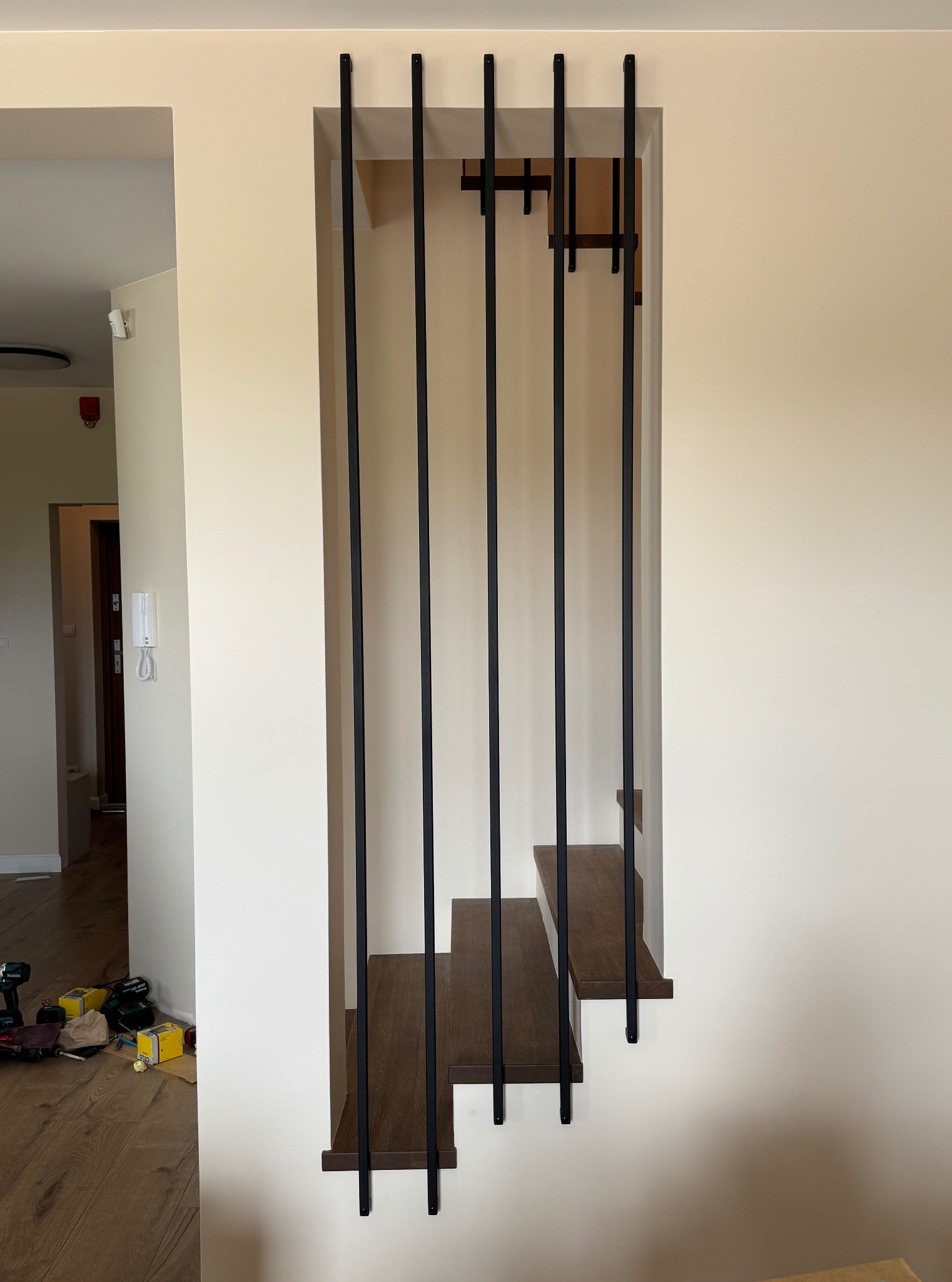 Nowoczesne schody z drewnianymi stopniami i minimalistyczną, czarną, metalową konstrukcją, wkomponowane w jasną ścianę. Widoczne narzędzia na podłodze.
