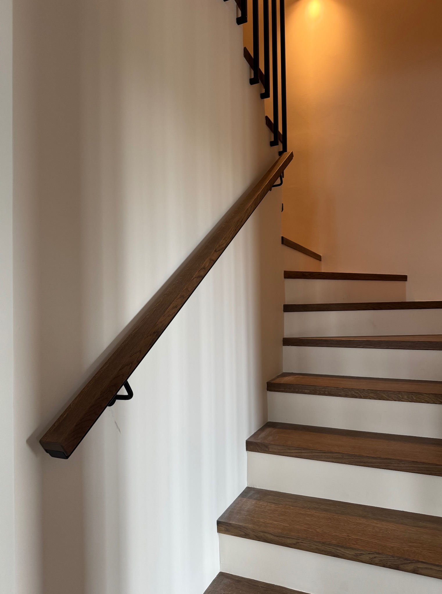 Drewniana poręcz schodowa z czarnymi uchwytami na białej ścianie, widoczne schody z drewnianymi stopniami i białą podstopnicą, czarne metalowe elementy balustrady.