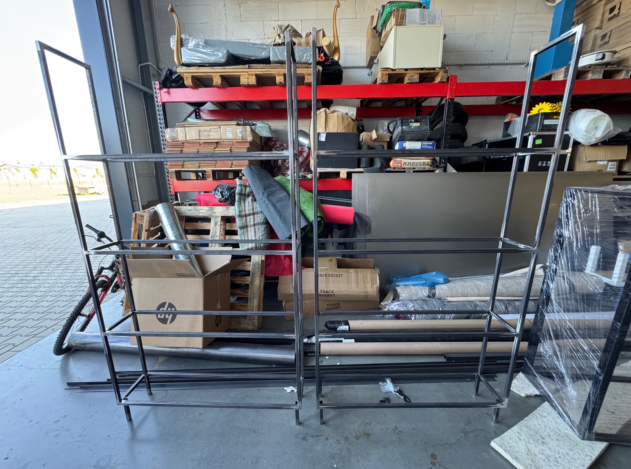 Surowe, metalowe konstrukcje regałów na tle magazynu z paletami i kartonami. Widoczne spawy, proste linie i minimalistyczny design.