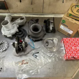 Rozłożony turbosprężarka samochodowa z częściami zamiennymi i akcesoriami na metalowym stole warsztatowym w serwisie samochodowym. Widoczne pudełka firm Melett i Elring.