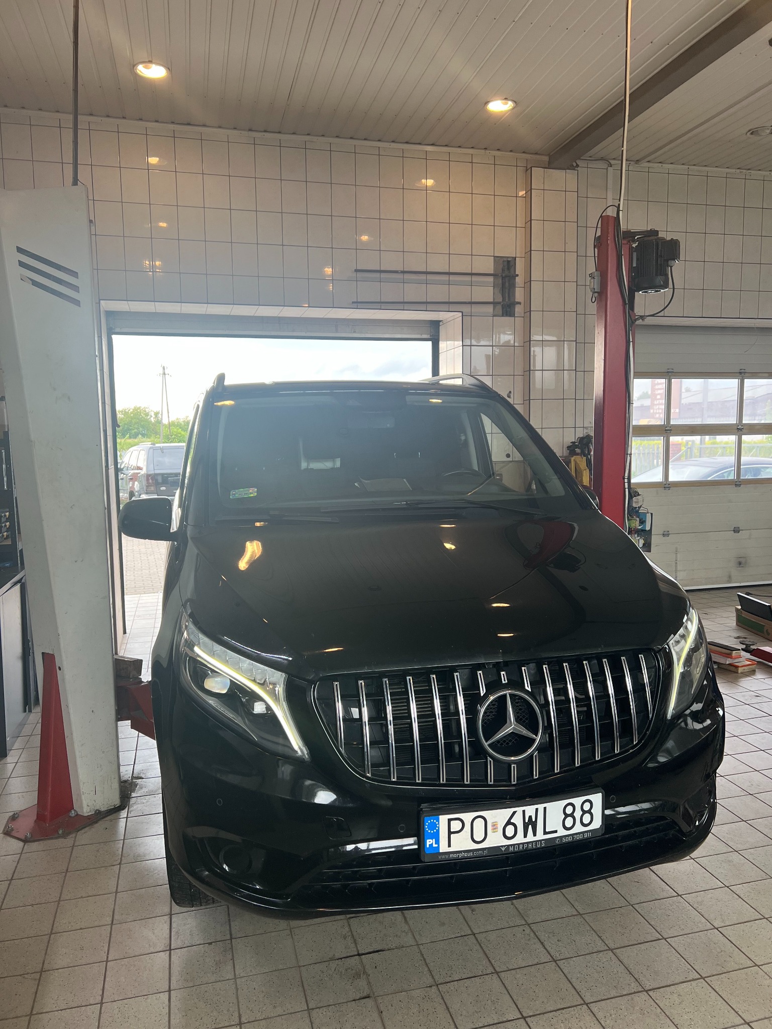 Czarny Mercedes V-klasa na podnośniku w warsztacie. Widoczna tablica rejestracyjna oraz element podnośnika. Tło stanowią białe płytki i brama wjazdowa.