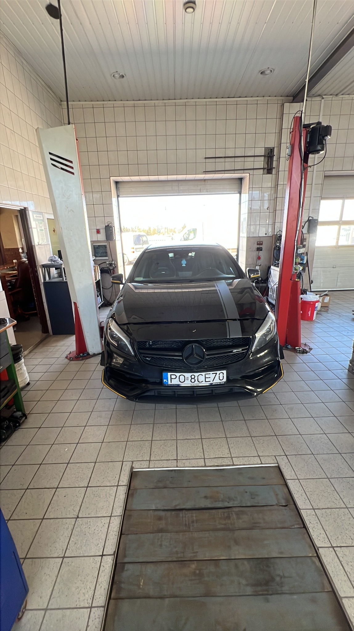 Czarny Mercedes A-klasa z szarym pasem na masce, zaparkowany w warsztacie z podnośnikiem i kanałem. Widok z przodu na tle otwartej bramy garażowej.