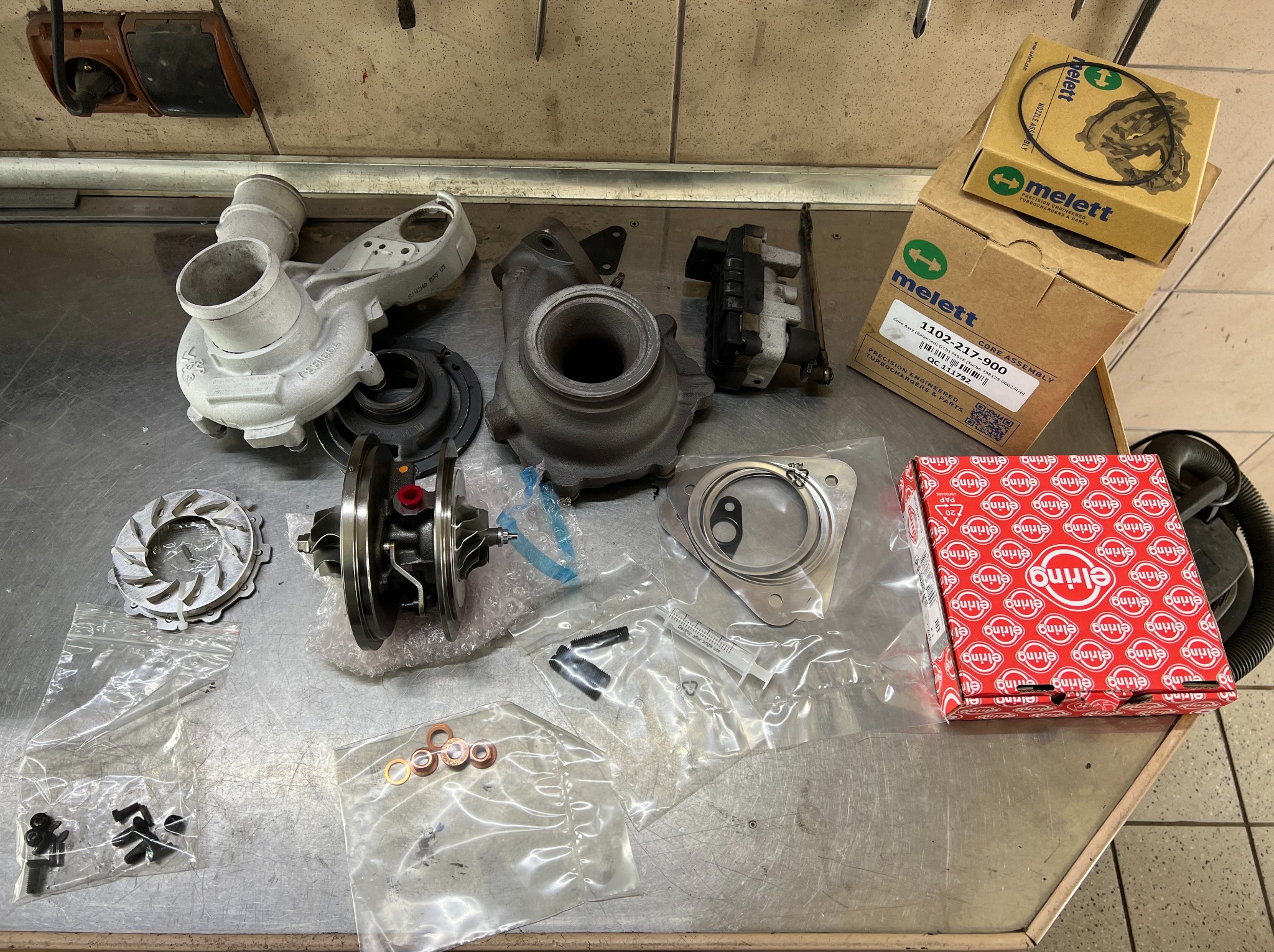 Rozłożony turbosprężarka samochodowa z częściami zamiennymi i akcesoriami na metalowym stole warsztatowym w serwisie samochodowym. Widoczne pudełka firm Melett i Elring.