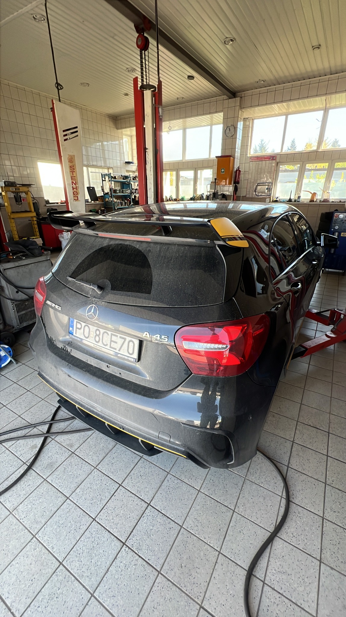 Czarny, lekko zakurzony Mercedes-Benz A45 AMG z żółtymi akcentami, widziany z tyłu w warsztacie samochodowym z podnośnikiem i narzędziami.