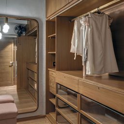 CONCEPT TEAM SPÓŁKA Z OGRANICZONĄ ODPOWIEDZIALNOŚCIĄ - Drewniana szafa wnękowa z lustrem i oświetleniem LED. Widoczne szklane półki i wieszaki z ubraniami. Ciepłe światło podkreśla naturalny odcień drewna.