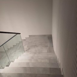 Usługi remontowe Dawid Soliński - Wykończone schody z szarych płytek, szklana balustrada z czarną ramą, dekoracyjna ściana z pionowymi żłobieniami. Minimalistyczny design.