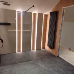 Nowoczesna łazienka z kabiną prysznicową typu walk-in, grzejnikiem żeberkowym i podświetlanymi panelami ściennymi w geometryczny wzór jodełki.