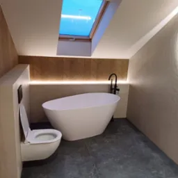 Nowoczesna łazienka na poddaszu z wolnostojącą wanną, toaletą podwieszaną i oknem dachowym, wykończona w jasnych barwach z akcentami drewna i podświetleniem LED.