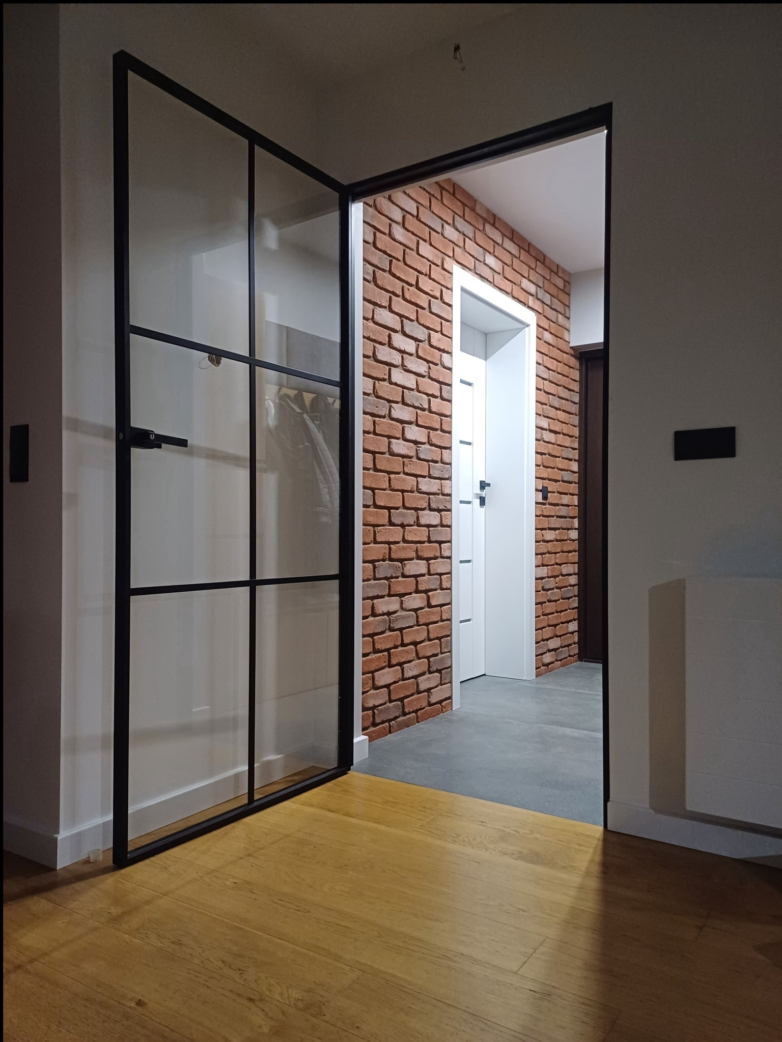 Nowoczesne wnętrze: przeszklone drzwi z czarnymi szprosami, ściana z czerwonej cegły, drewniana podłoga i białe drzwi w tle.