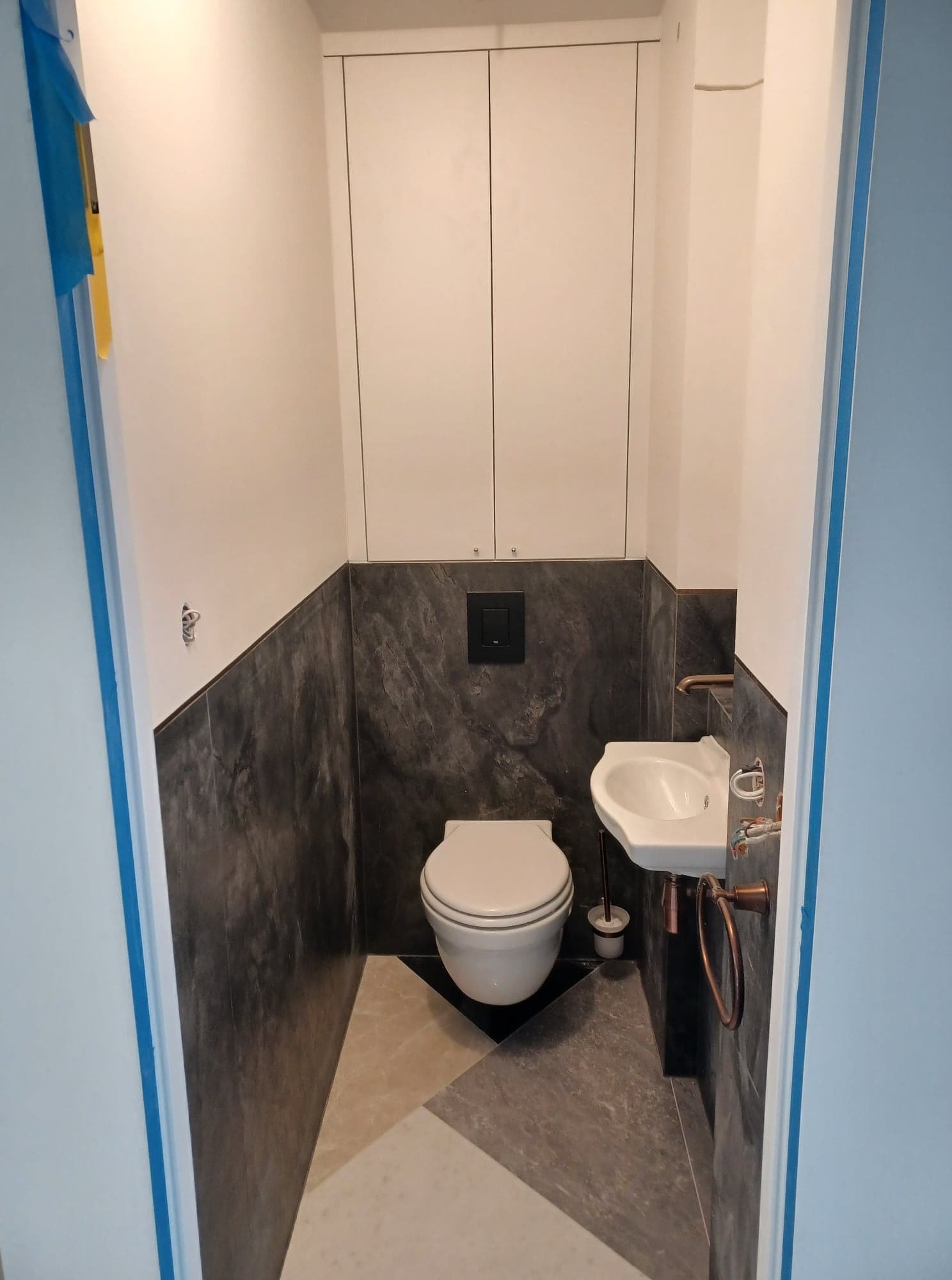 Nowoczesna toaleta po remoncie: ciemne płytki imitujące kamień na ścianach, biała armatura i szafka nad toaletą. Geometryczny wzór na podłodze.