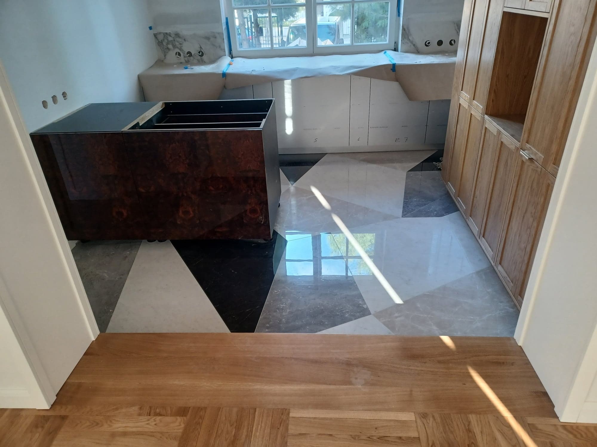 Wnętrze w trakcie prac: marmurowe płytki podłogowe w geometryczny wzór, zabudowa okienna zabezpieczona folią i drewniana szafa wnękowa. Widok z sąsiedniego pomieszczenia.