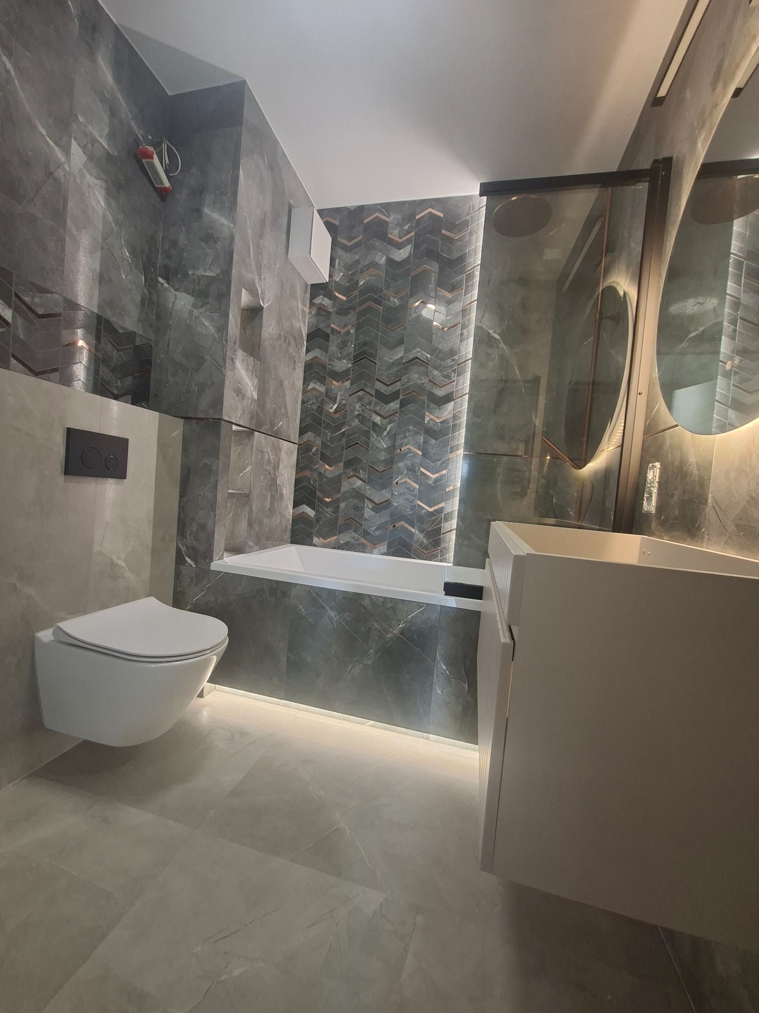 Nowoczesna łazienka w szarościach z podświetleniem LED: wisząca toaleta, wanna i umywalka. Na ścianach płytki imitujące marmur oraz dekoracyjne w jodełkę.