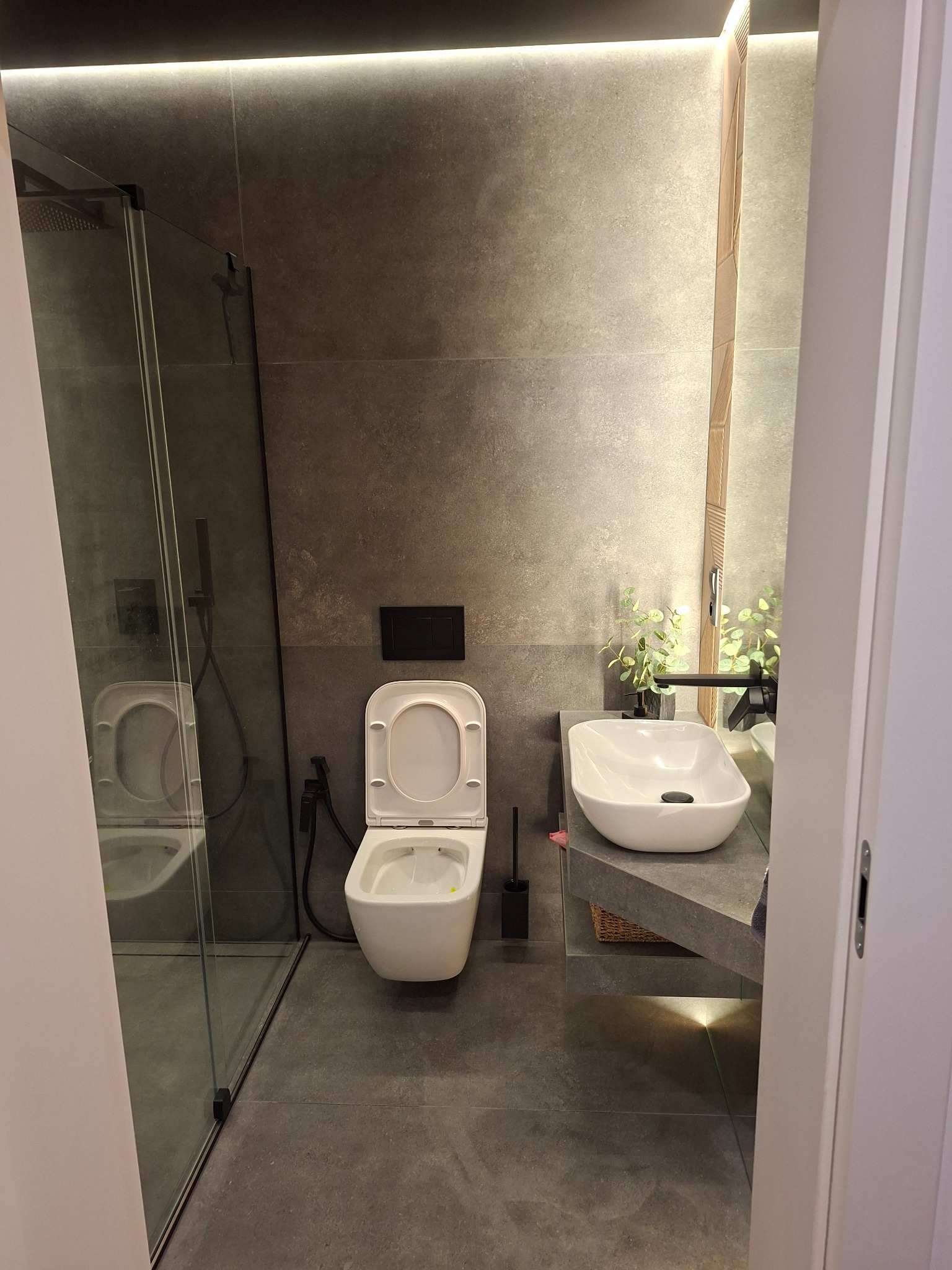 Nowoczesna łazienka w szarościach: podwieszana toaleta z otwartą klapą, przeszklony prysznic, umywalka nablatowa i subtelne oświetlenie LED.