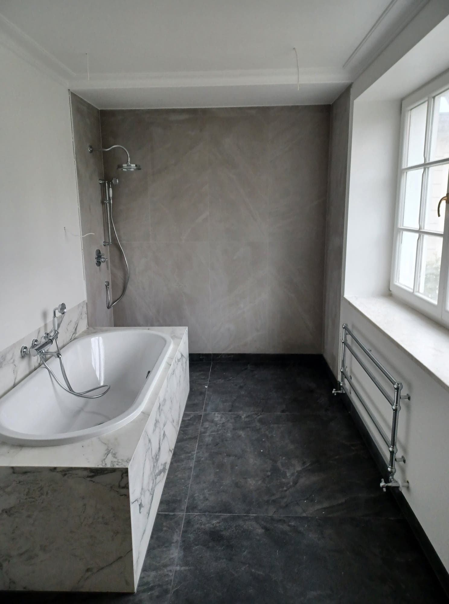 Nowoczesna łazienka z marmurową obudową wanny, szarymi płytkami ściennymi i podłogowymi, oraz grzejnikiem drabinkowym pod oknem. Minimalistyczny design.