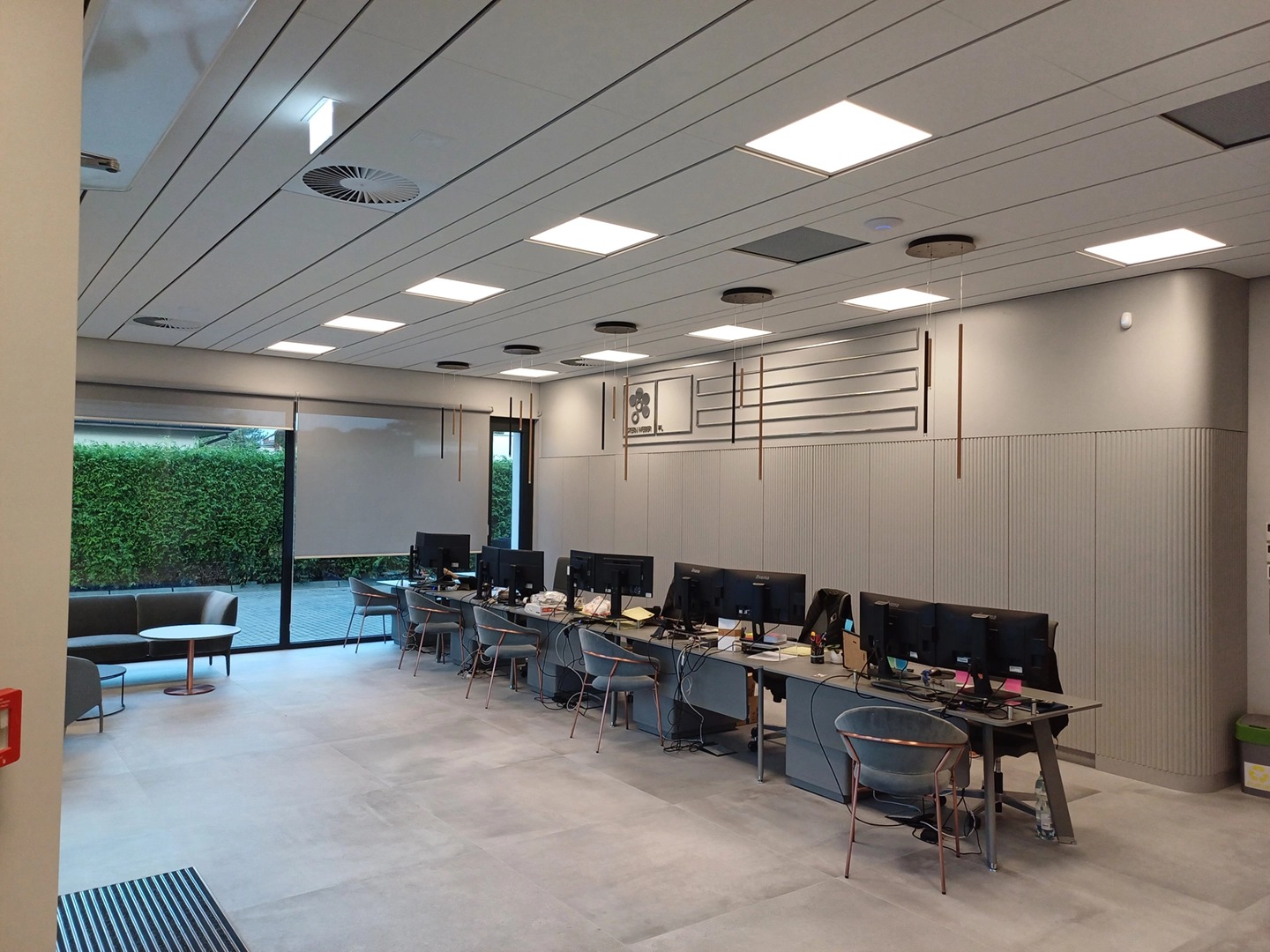 Jasne biuro po remoncie: szare ściany z dekoracyjnymi listwami, rzędy stanowisk komputerowych, duże okno z widokiem na zieleń. Nowoczesne oświetlenie sufitowe.