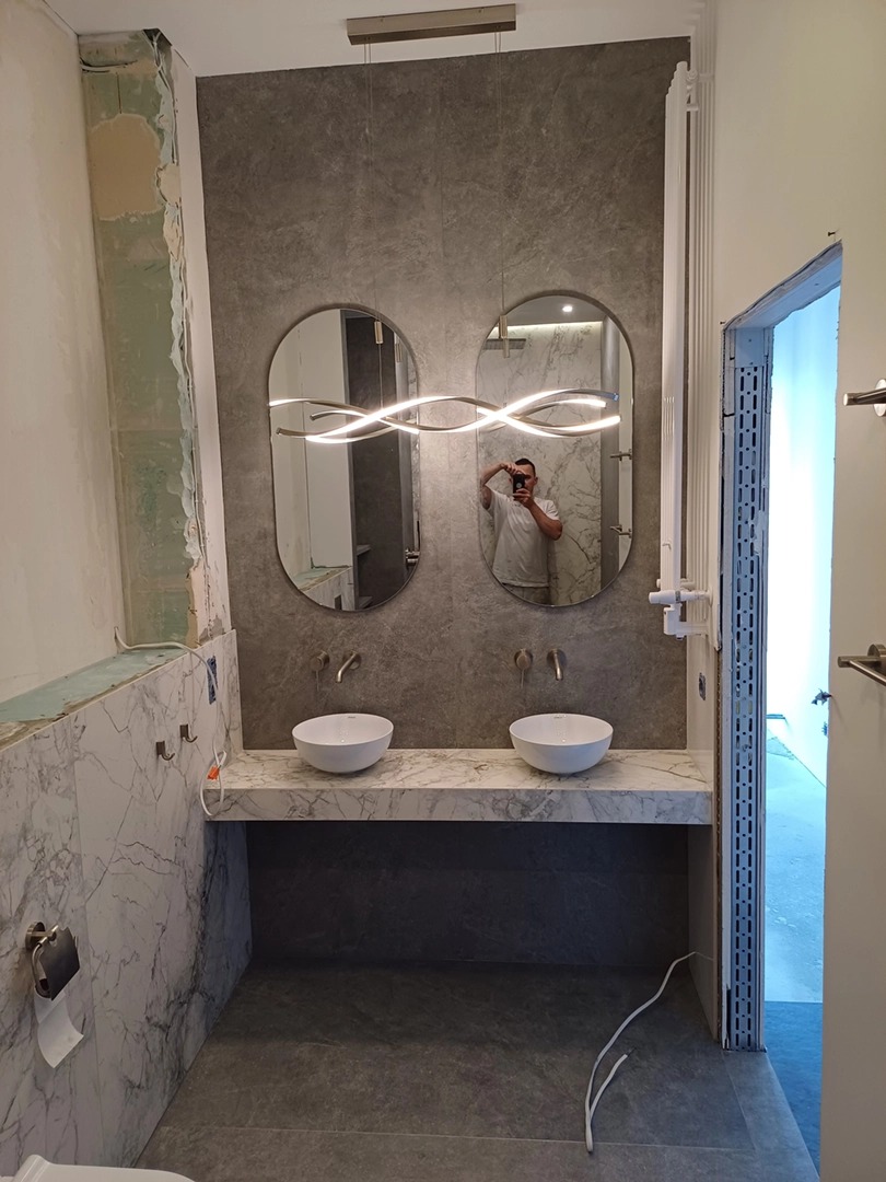 Nowoczesna łazienka w trakcie prac: marmurowe płytki, podwójna umywalka, lustra owalne z oświetleniem LED. Widoczne niedokończone wykończenia.