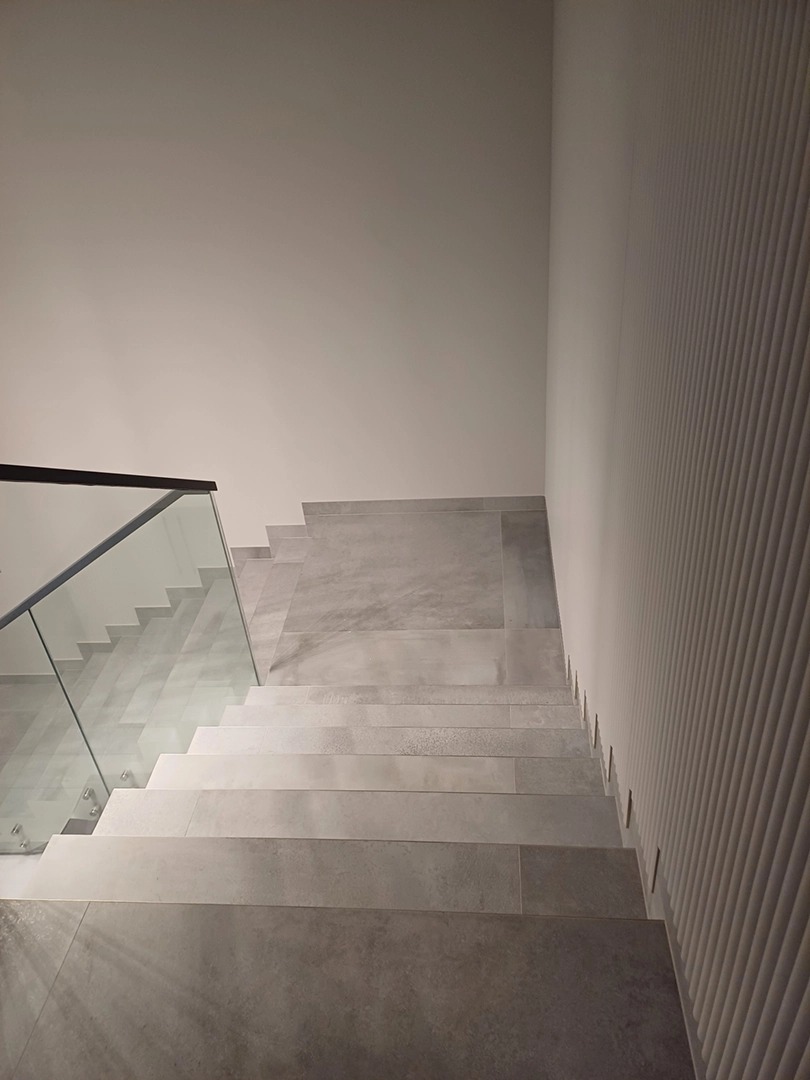 Wykończone schody z szarych płytek, szklana balustrada z czarną ramą, dekoracyjna ściana z pionowymi żłobieniami. Minimalistyczny design.