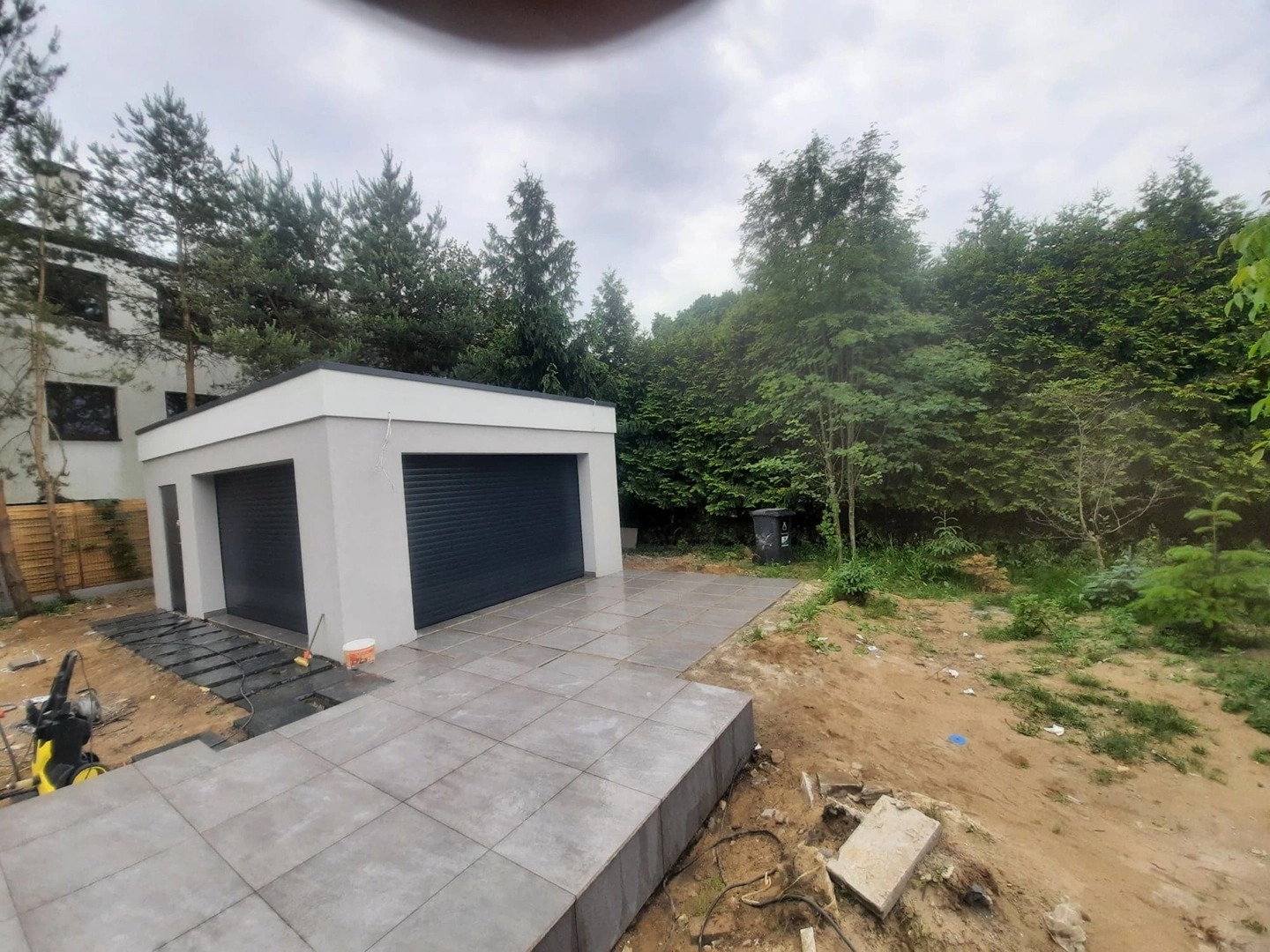 Nowoczesny, jasnoszary garaż z dwiema bramami rolowanymi, z ułożoną kostką brukową na podjeździe, otoczony zielenią i drzewami, w pochmurny dzień.