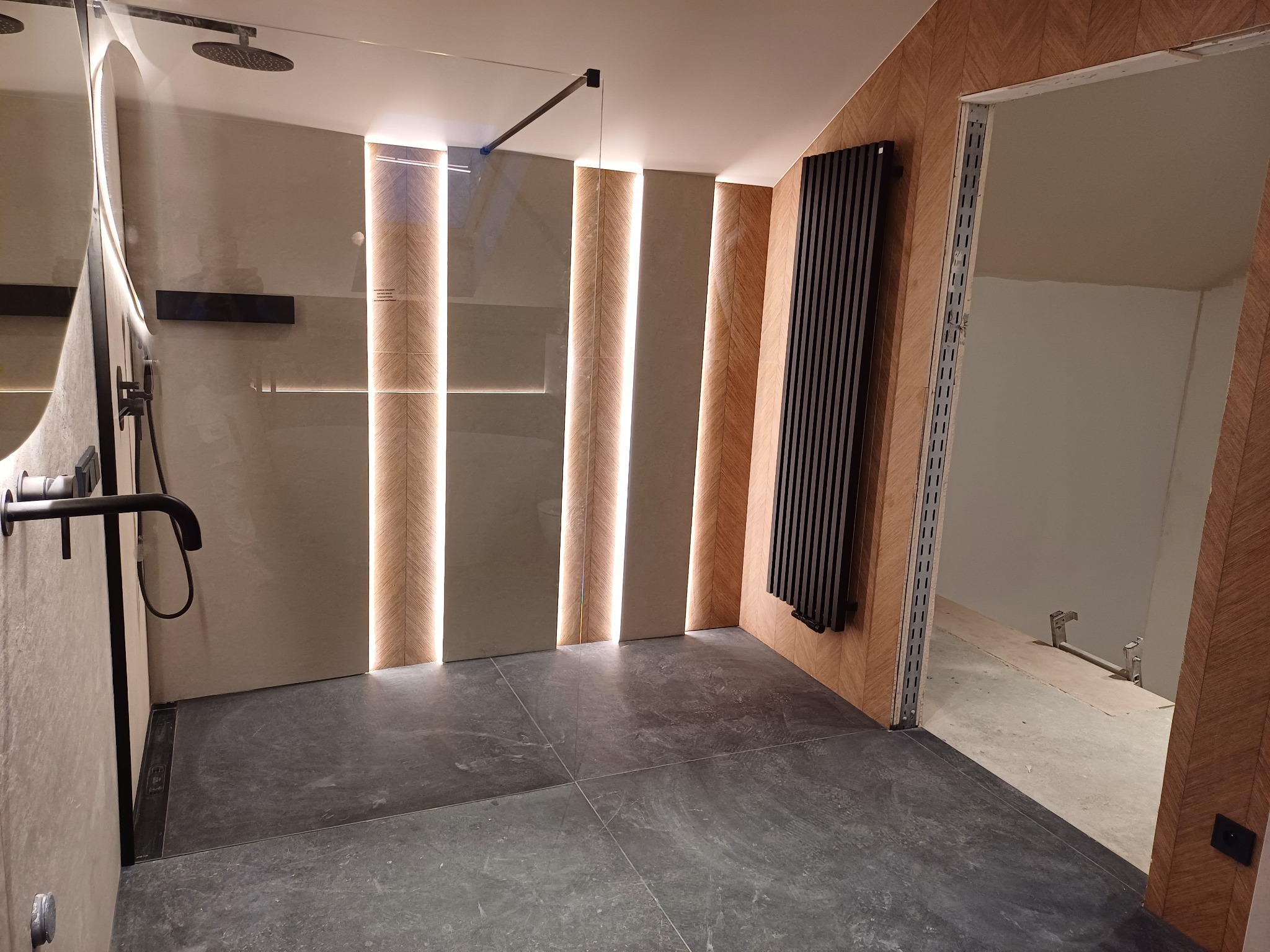 Nowoczesna łazienka z kabiną prysznicową typu walk-in, grzejnikiem żeberkowym i podświetlanymi panelami ściennymi w geometryczny wzór jodełki.