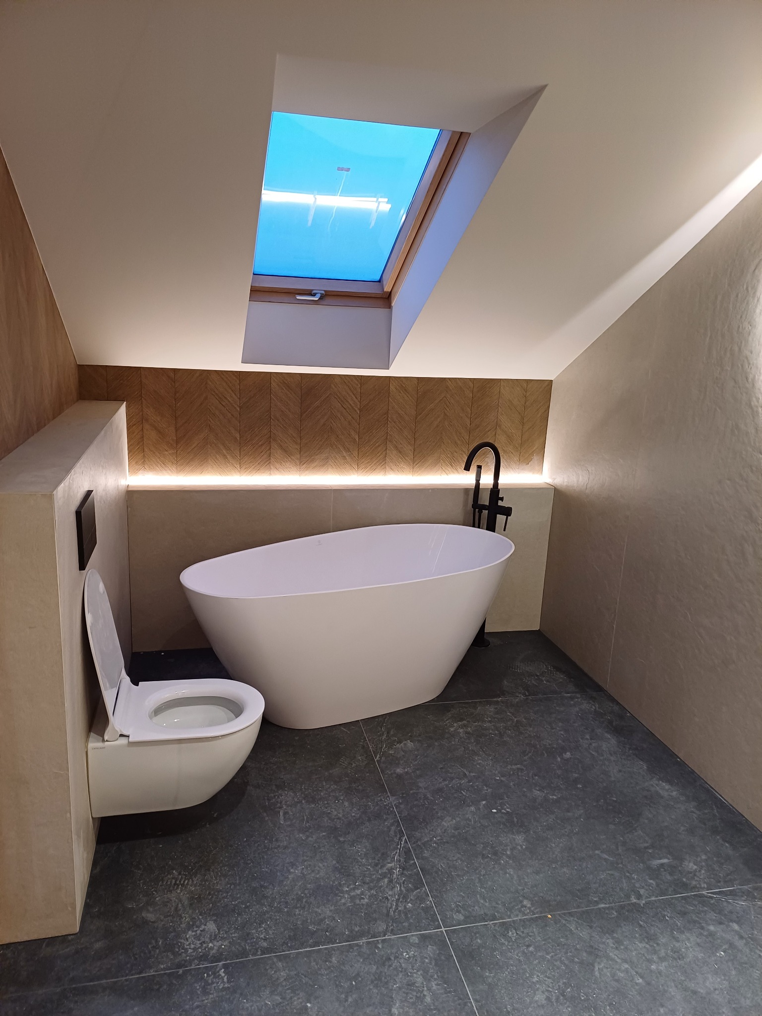 Nowoczesna łazienka na poddaszu z wolnostojącą wanną, toaletą podwieszaną i oknem dachowym, wykończona w jasnych barwach z akcentami drewna i podświetleniem LED.