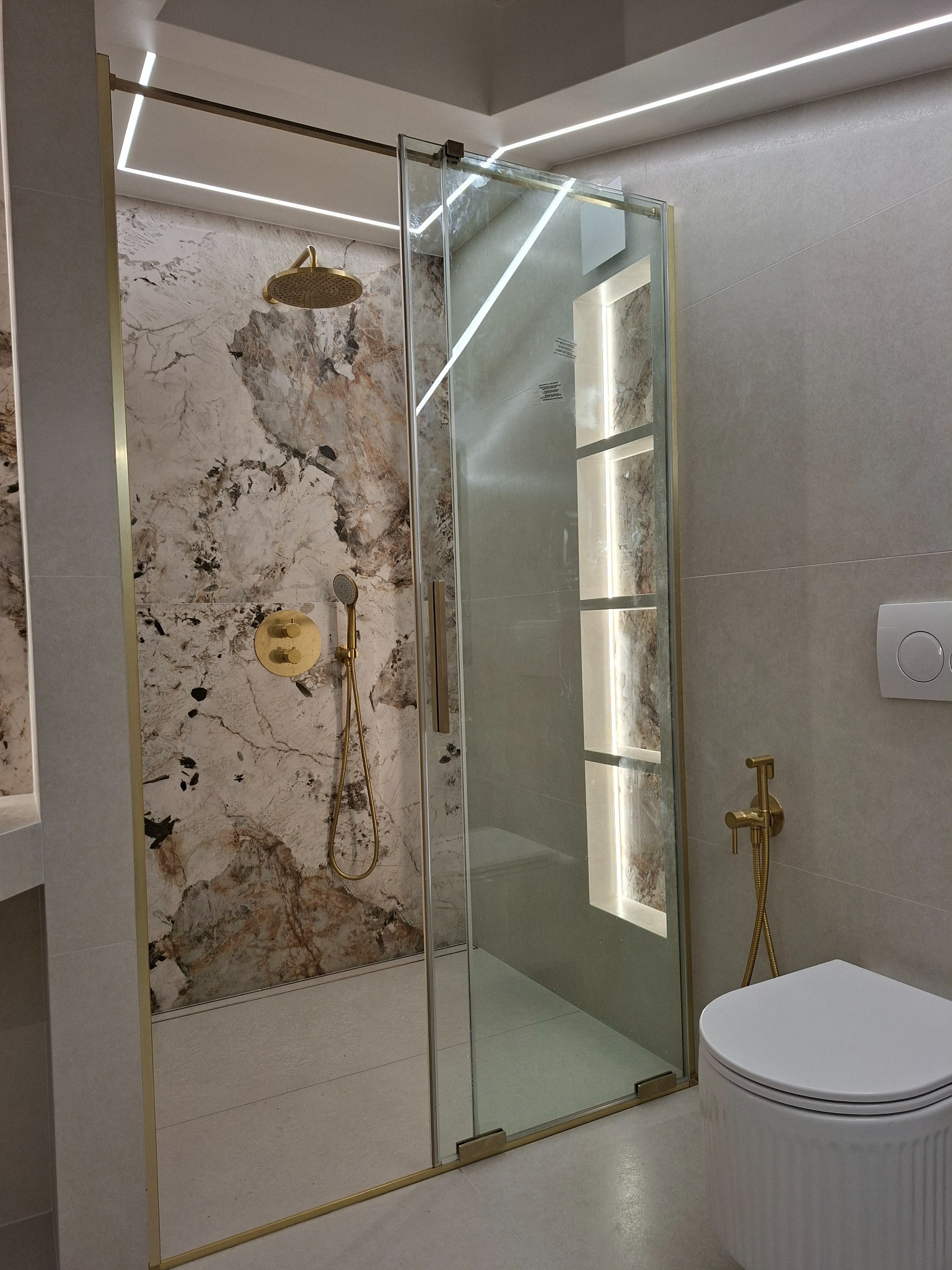 Nowoczesna łazienka z kabiną prysznicową typu walk-in, złotą armaturą i podświetlanymi niszami w ścianie. Marmurowa ściana pod prysznicem dodaje elegancji.