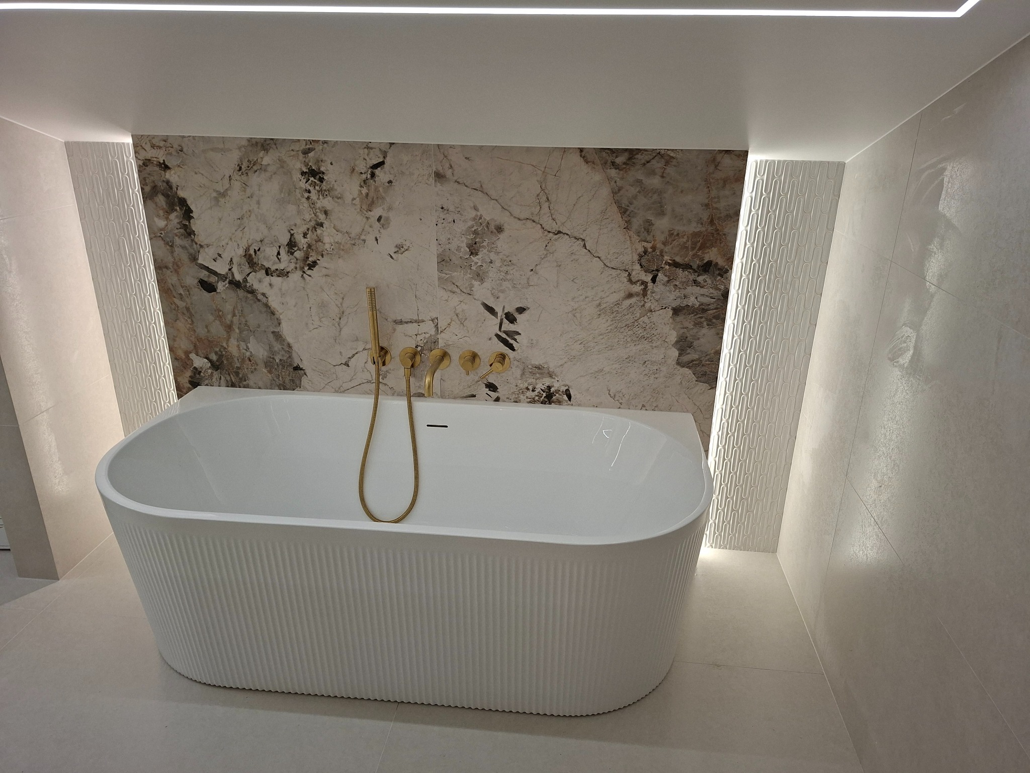 Nowoczesna łazienka z wolnostojącą wanną o ryflowanej powierzchni, złota armatura na tle marmurowej ściany, oświetlenie LED wzdłuż ścian.