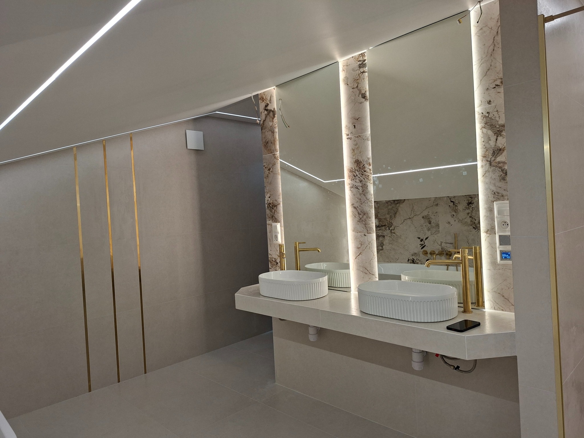 Nowoczesna łazienka z dwoma umywalkami nablatowymi, złotą armaturą i podświetlanymi lustrami w marmurowych obudowach. Minimalistyczny design z akcentami złota.